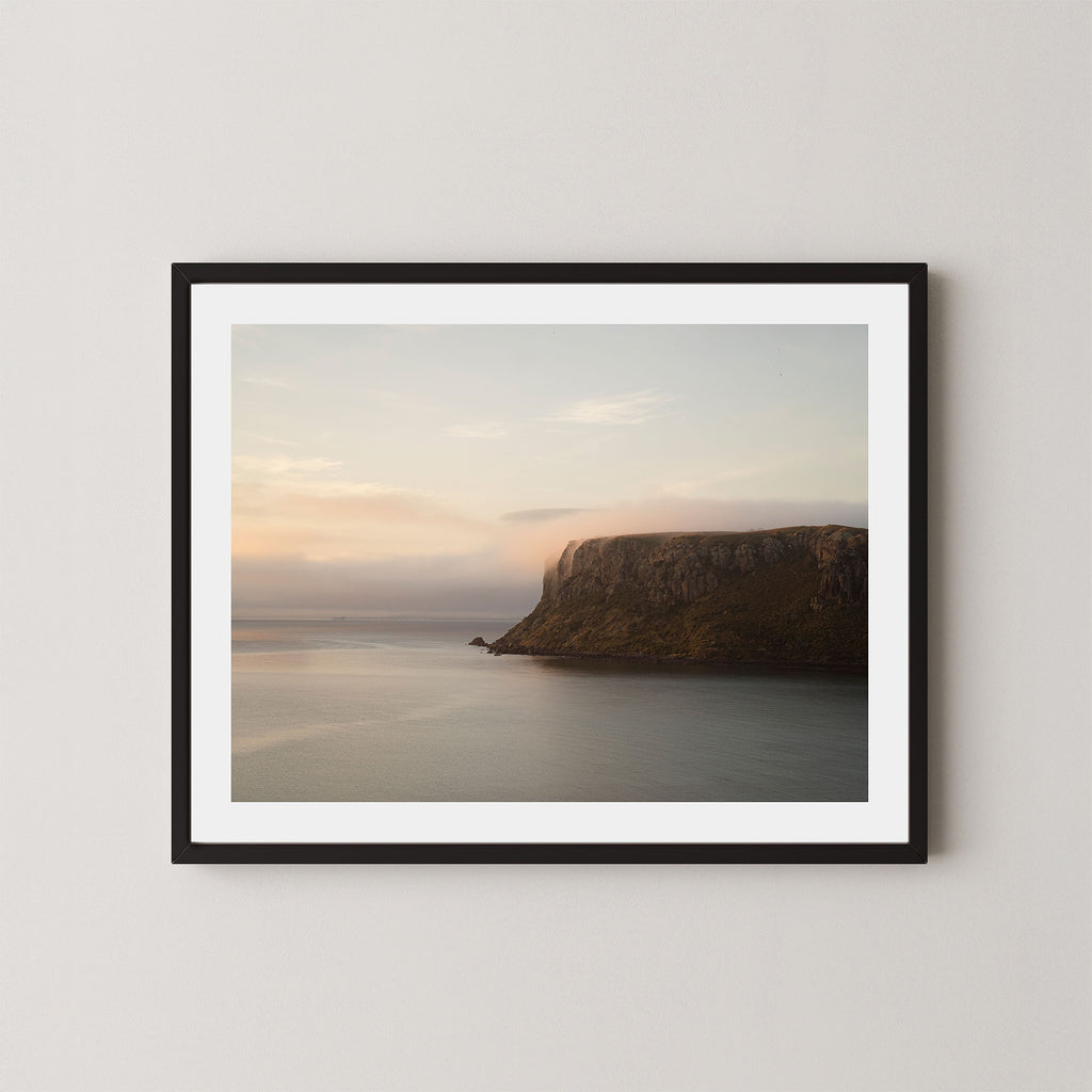 Silent Coast art print of a coastal cliff and sunrise in Stanley, Tasmania, displayed on a white wall in a black frame.