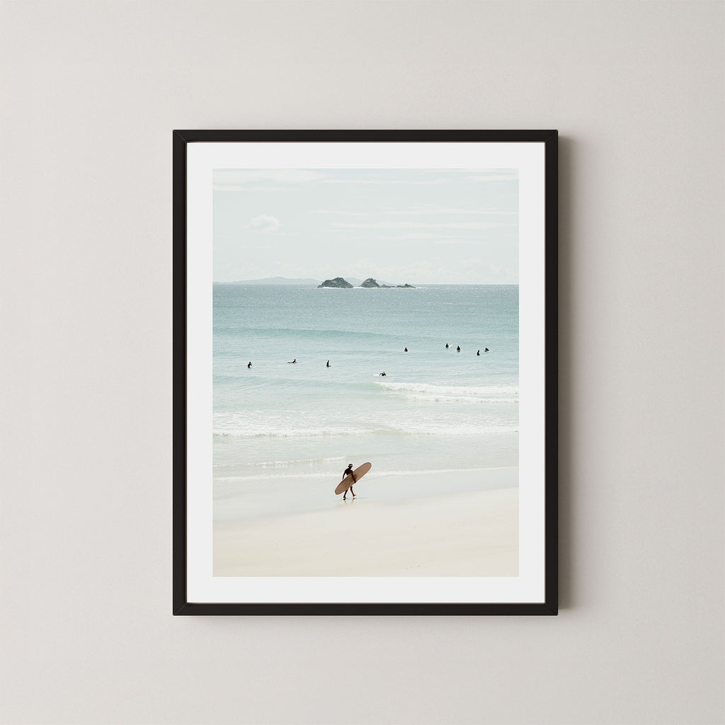 Wategos Calm art print of a surfer walking with a surfboard on the beach in Byron Bay, Australia, with the ocean and other surfers in the background, displayed on a wall in a black frame.