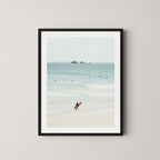 Wategos Calm art print of a surfer walking with a surfboard on the beach in Byron Bay, Australia, with the ocean and other surfers in the background, displayed on a wall in a black frame.
