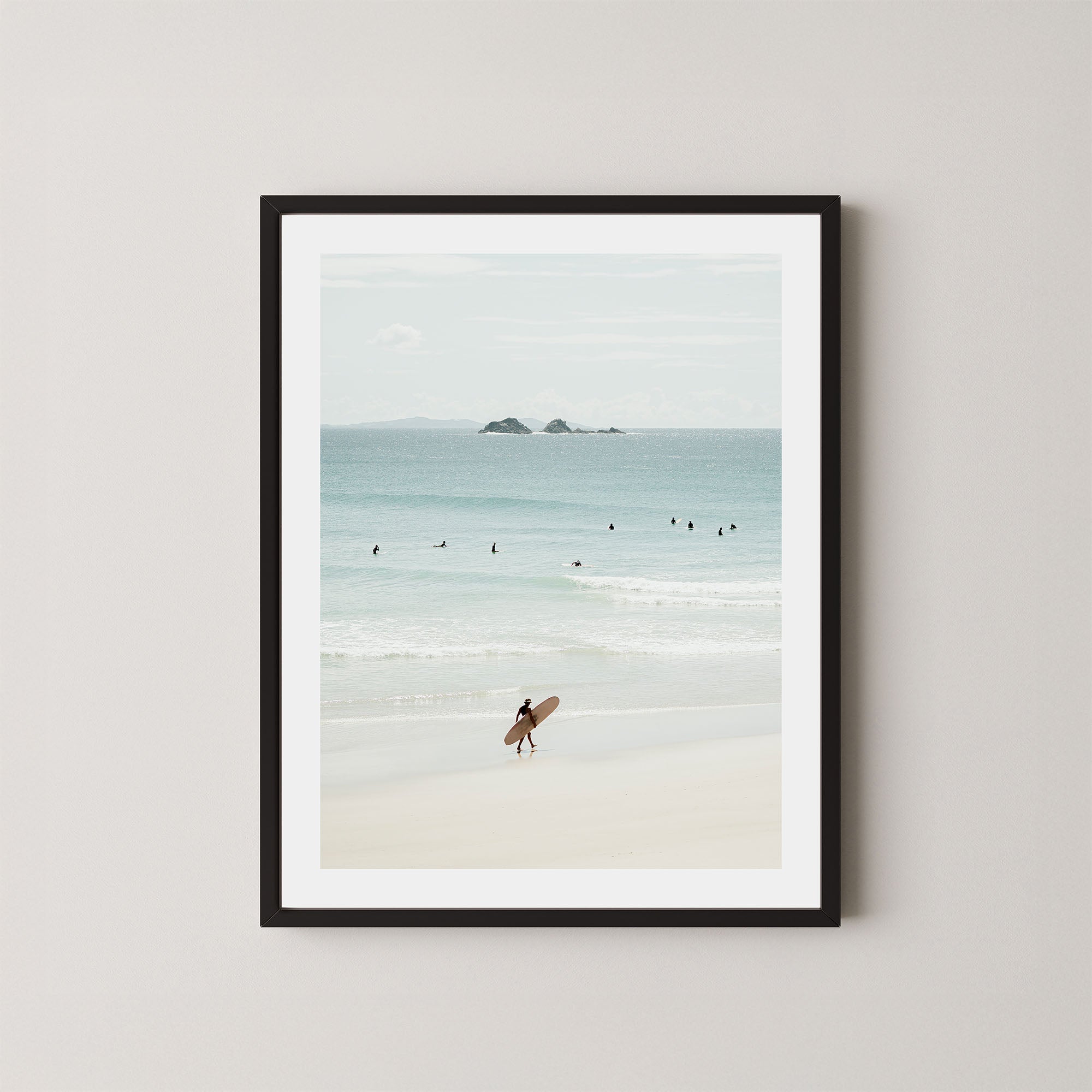 Wategos Calm art print of a surfer walking with a surfboard on the beach in Byron Bay, Australia, with the ocean and other surfers in the background, displayed on a wall in a black frame.