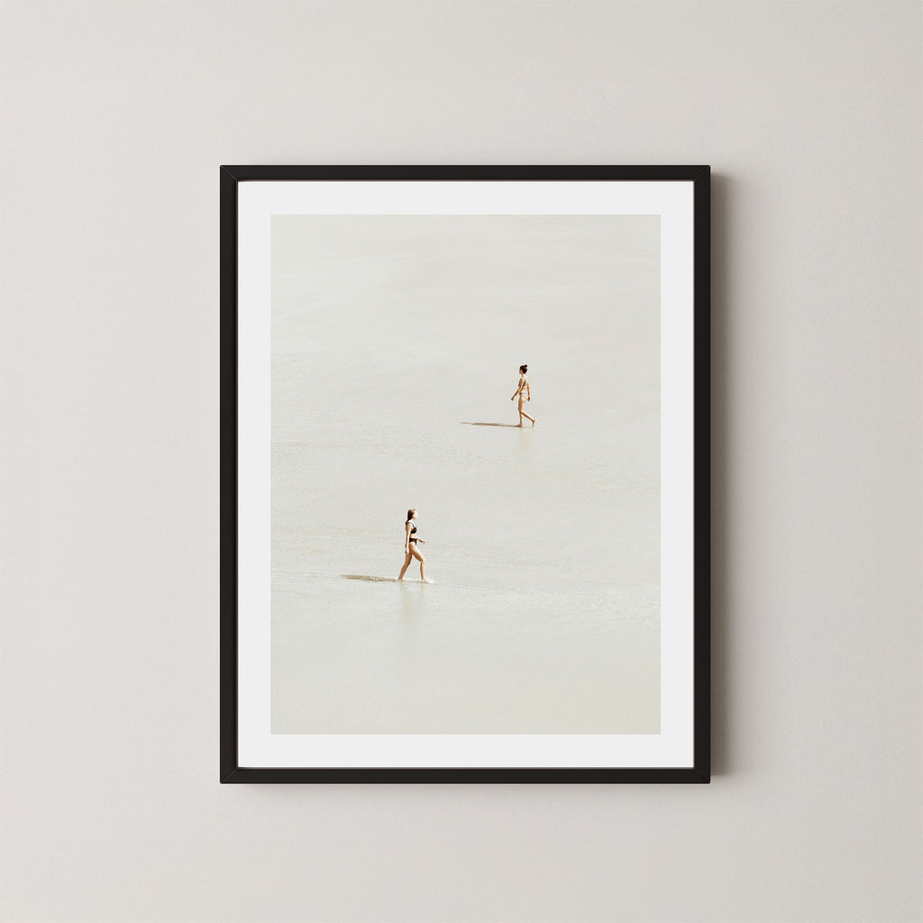 Beach Crossing art print featuring two women walking in opposite directions on a light, sandy beach at low tide in Byron Bay, Australia, displayed on a wall in a black frame.