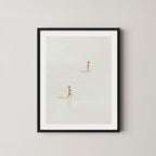 Beach Crossing art print featuring two women walking in opposite directions on a light, sandy beach at low tide in Byron Bay, Australia, displayed on a wall in a black frame.