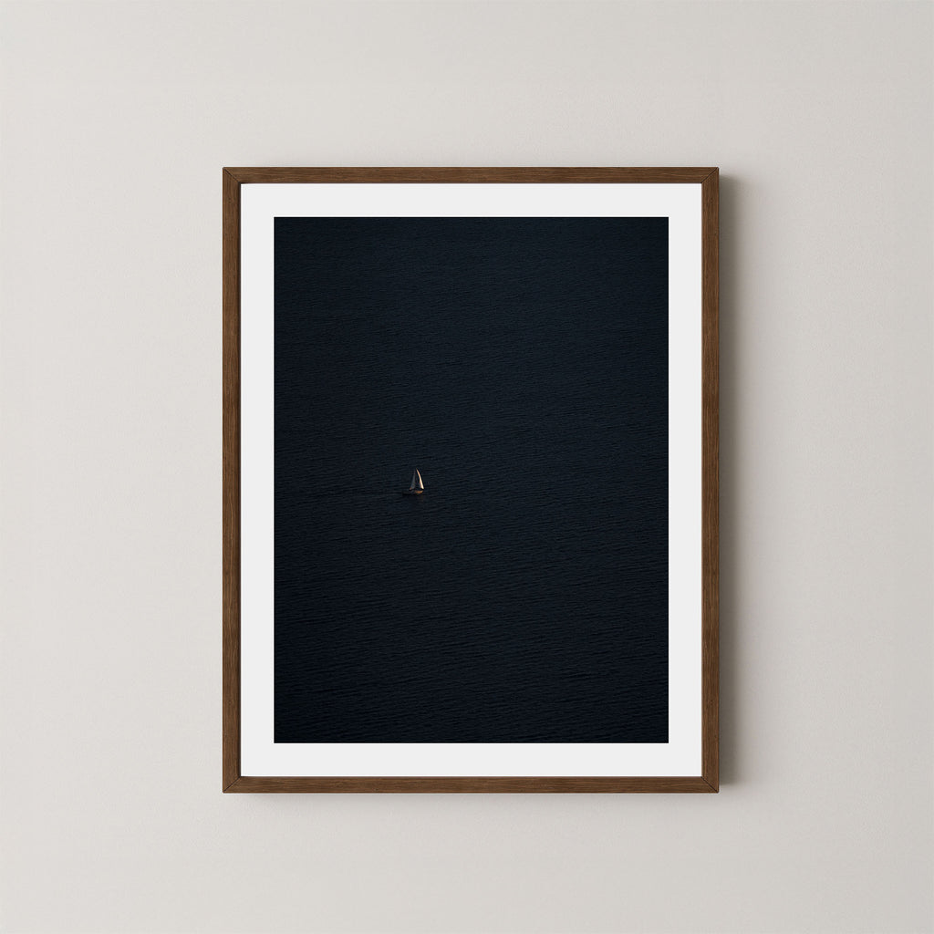 Voyage art print featuring a single sailing boat on the ocean, displayed on a wall in a dark walnut wooden frame.