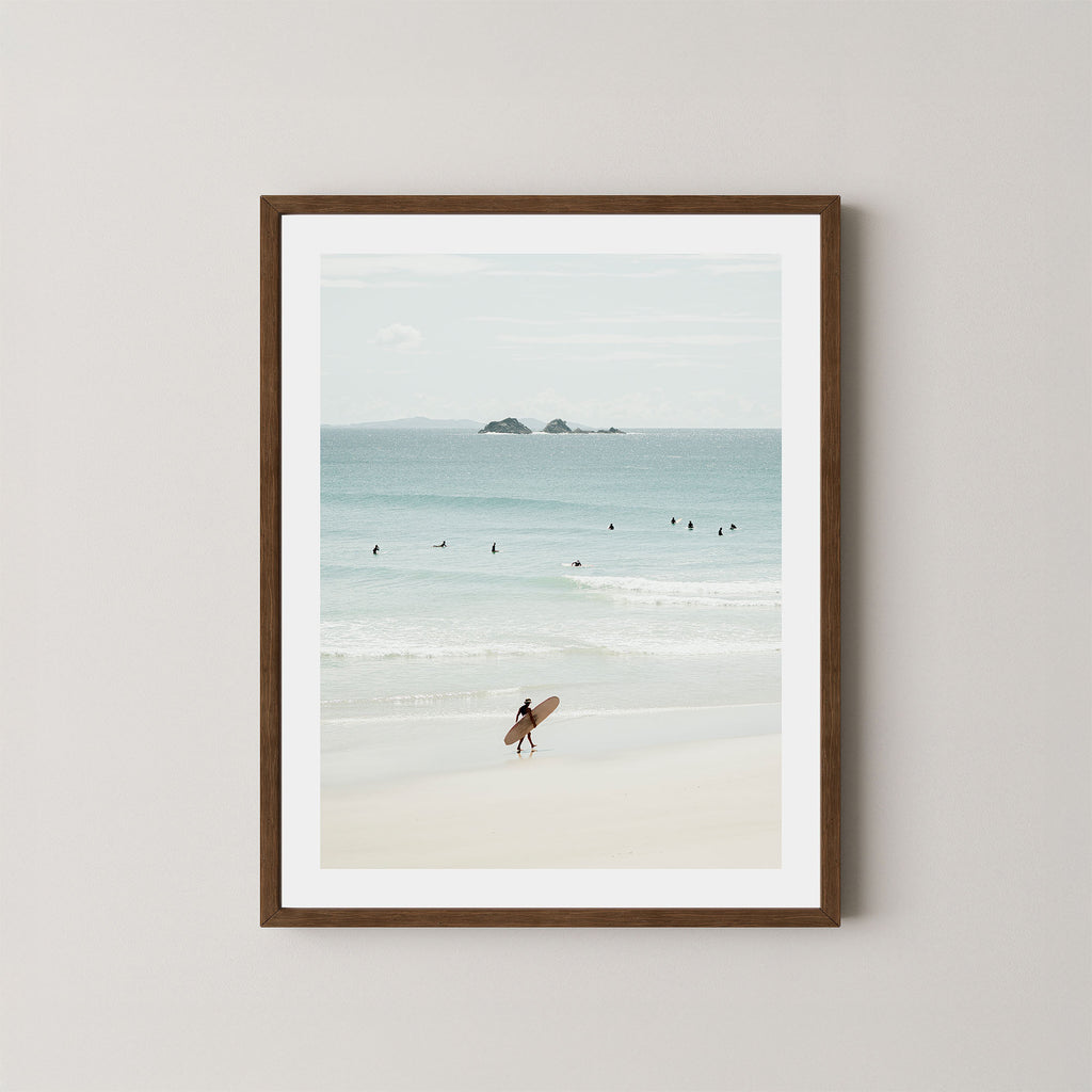 Wategos Calm art print of a surfer walking with a surfboard on the beach in Byron Bay, Australia, with the ocean and other surfers in the background, displayed on a wall in a dark wooden frame.