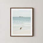 Wategos Calm art print of a surfer walking with a surfboard on the beach in Byron Bay, Australia, with the ocean and other surfers in the background, displayed on a wall in a dark wooden frame.