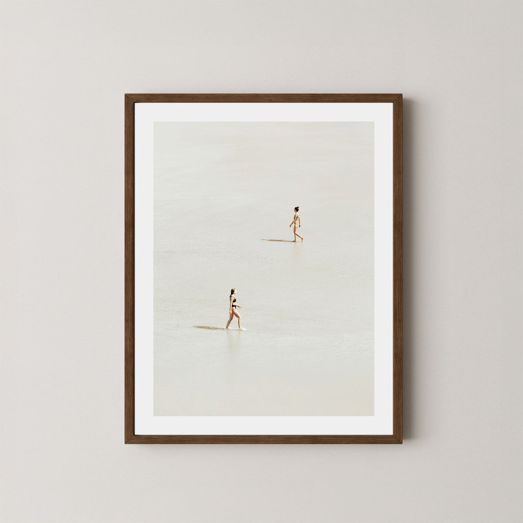 Beach Crossing art print featuring two women walking in opposite directions on a light, sandy beach at low tide in Byron Bay, Australia, displayed on a wall in a dark wooden frame.