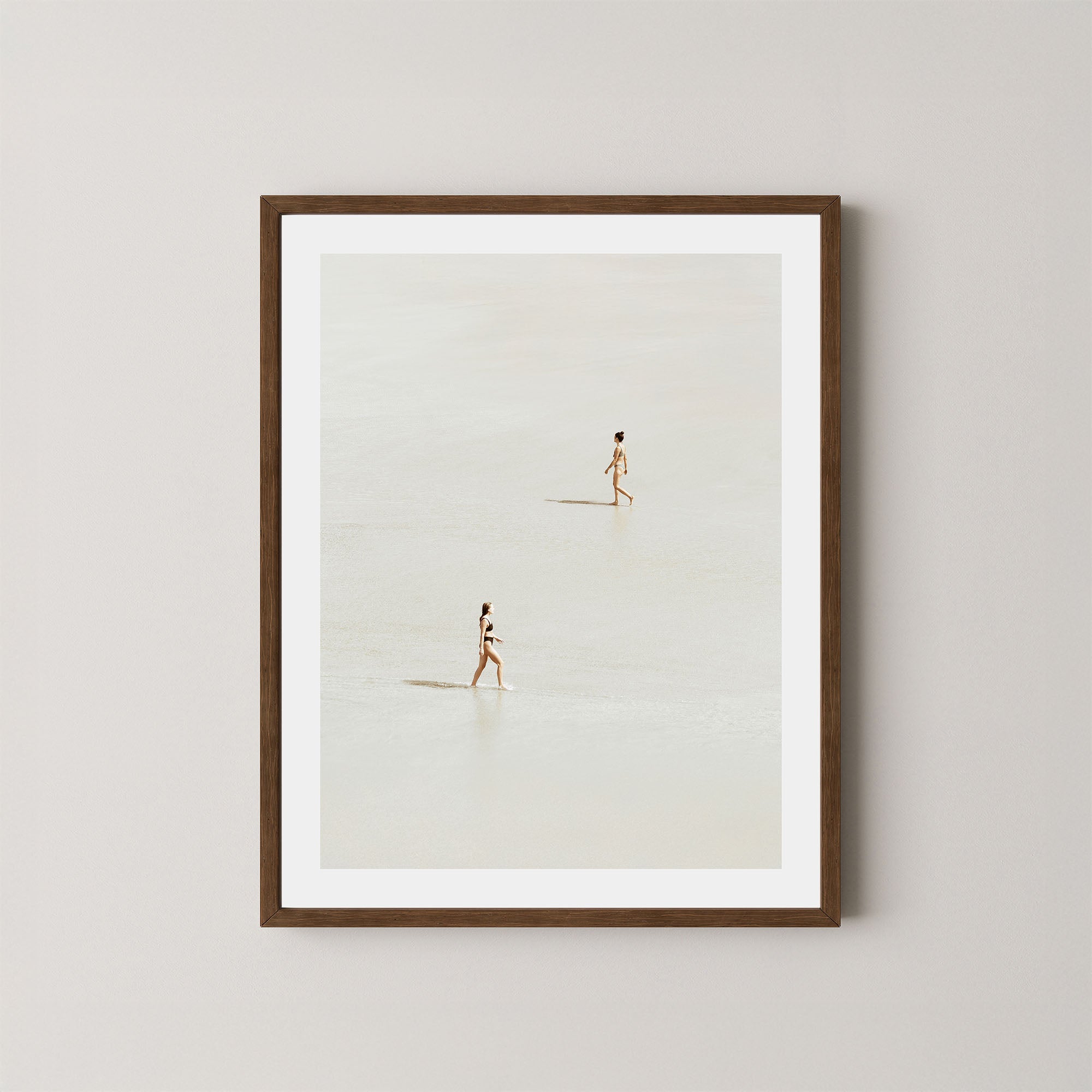 Beach Crossing art print featuring two women walking in opposite directions on a light, sandy beach at low tide in Byron Bay, Australia, displayed on a wall in a dark wooden frame.