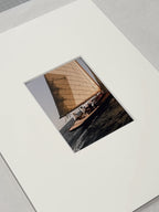 A square photographic print of a sailboat on dark water, mounted or presented on a large white mat against a light gray background.