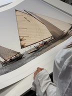 A large framed art print depicting two classic sailing yachts on the water, shown being held by a person wearing a white shirt.