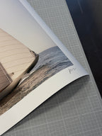 A close-up of a printed photographic art piece showing part of a boat and water, laid on a gray gridded cutting mat. The print has a white border and a small handwritten signature near the lower right edge.