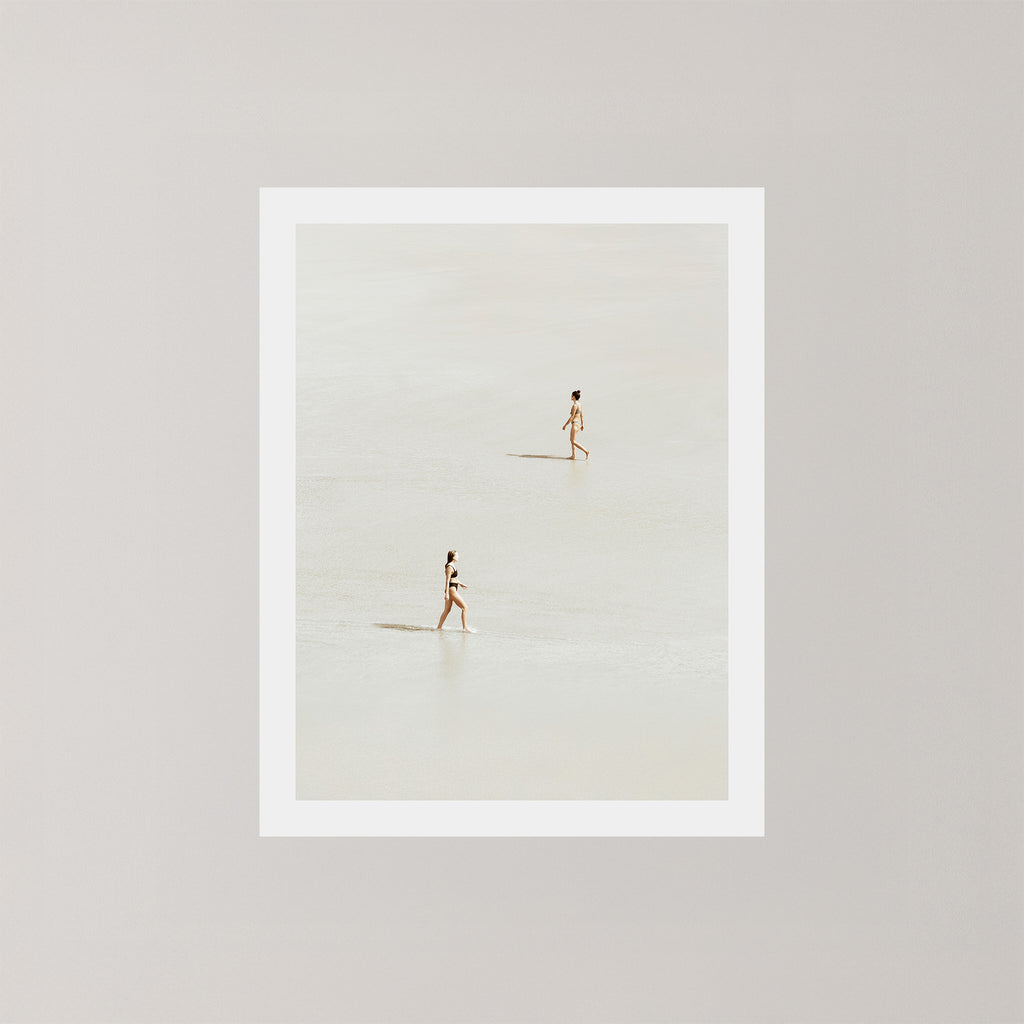 Beach Crossing photography art print featuring two women walking in opposite directions on a light, sandy beach at low tide in Byron Bay, Australia.