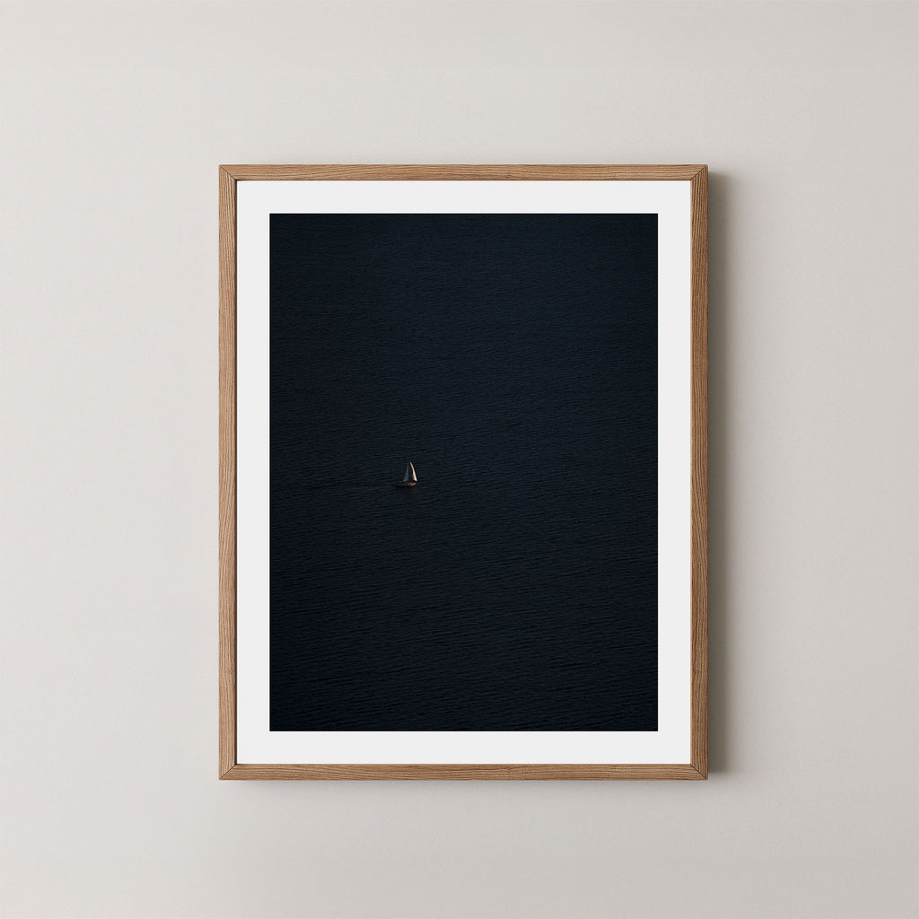 Voyage art print featuring a single sailing boat on the ocean, displayed on a wall in a natural wood oak frame.