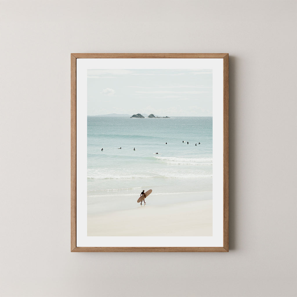 Wategos Calm art print of a surfer walking with a surfboard on the beach in Byron Bay, Australia, with the ocean and other surfers in the background, displayed on a wall in a natural wooden frame.