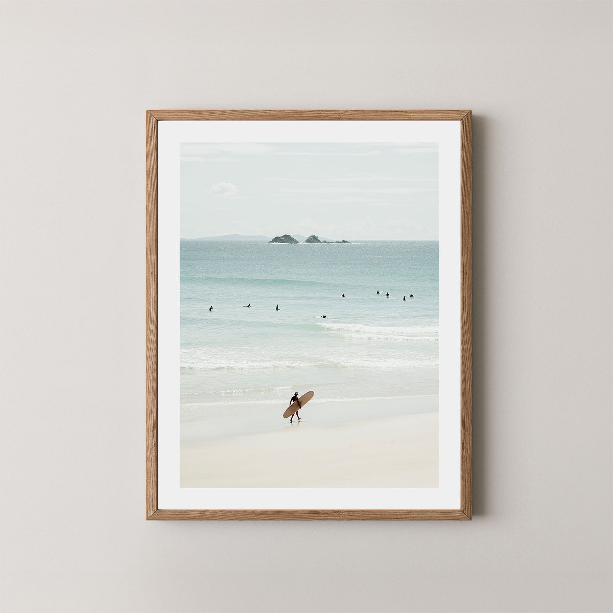 Wategos Calm art print of a surfer walking with a surfboard on the beach in Byron Bay, Australia, with the ocean and other surfers in the background, displayed on a wall in a natural wooden frame.