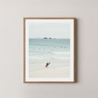 Wategos Calm art print of a surfer walking with a surfboard on the beach in Byron Bay, Australia, with the ocean and other surfers in the background, displayed on a wall in a natural wooden frame.