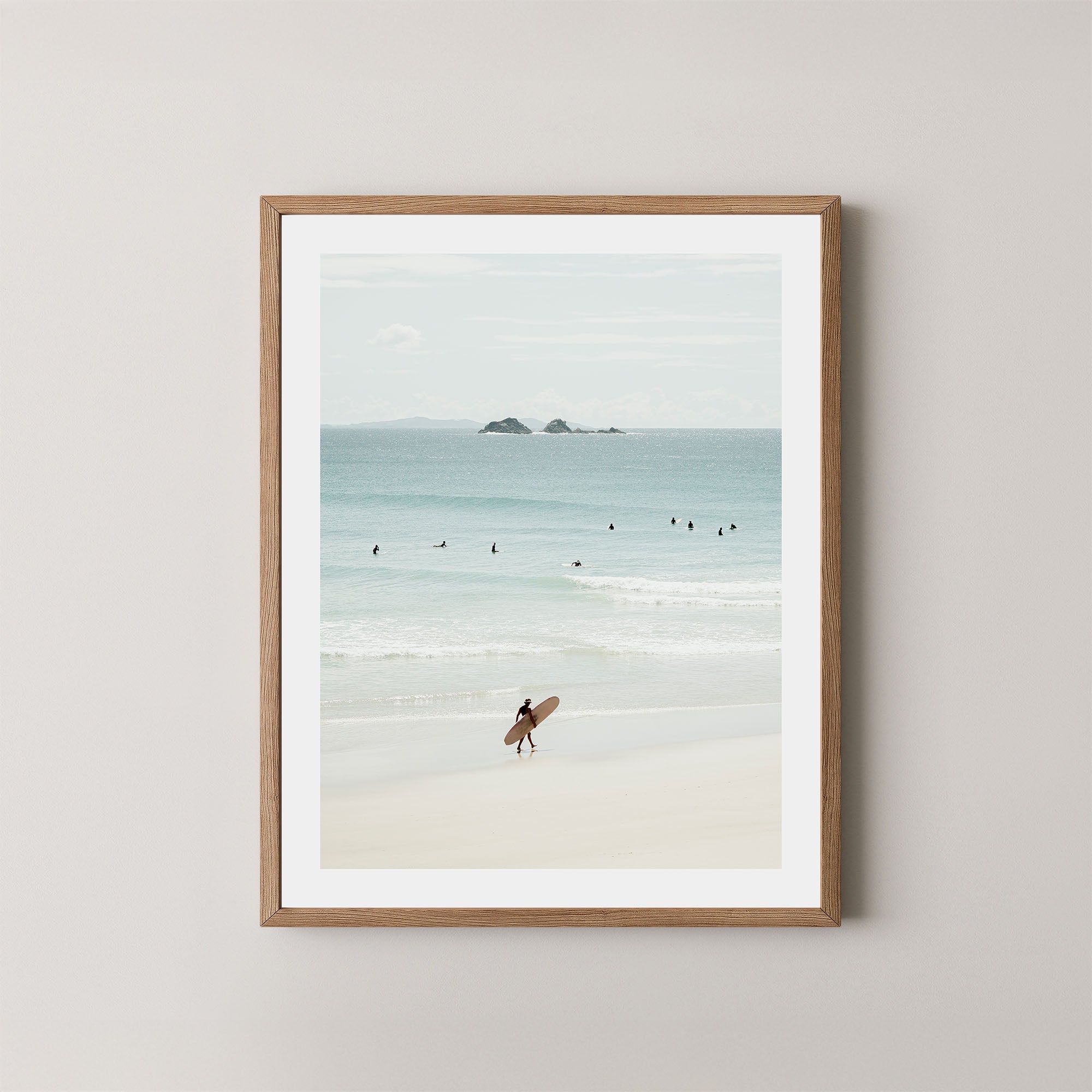 Wategos Calm art print of a surfer walking with a surfboard on the beach in Byron Bay, Australia, with the ocean and other surfers in the background, displayed on a wall in a natural wooden frame.