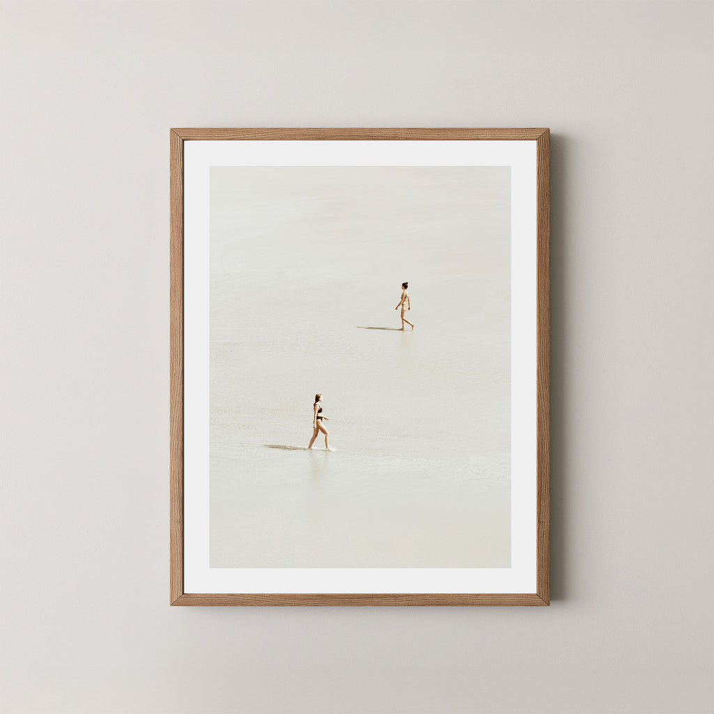 Beach Crossing art print featuring two women walking in opposite directions on a light, sandy beach at low tide in Byron Bay, Australia, displayed on a wall in a light wooden frame.