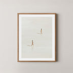 Beach Crossing art print featuring two women walking in opposite directions on a light, sandy beach at low tide in Byron Bay, Australia, displayed on a wall in a light wooden frame.