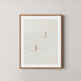 Beach Crossing art print featuring two women walking in opposite directions on a light, sandy beach at low tide in Byron Bay, Australia, displayed on a wall in a light wooden frame.