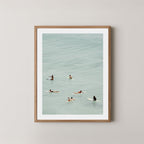 Fine photography artwork of people on surfboards in the water, displayed in a natural wooden frame. 