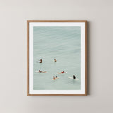 Fine photography artwork of people on surfboards in the water, displayed in a natural wooden frame. 