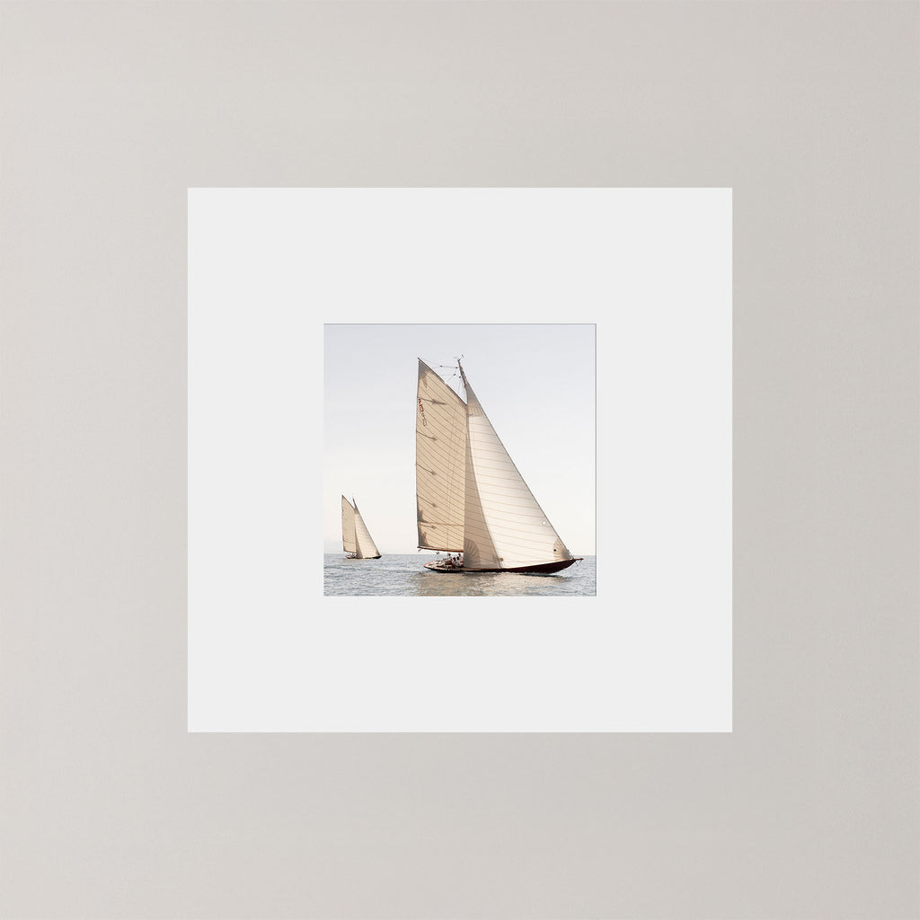 A square art print featuring a photograph of two sailboats on calm water under a pale sky, surrounded by a wide white mat on a light gray background.