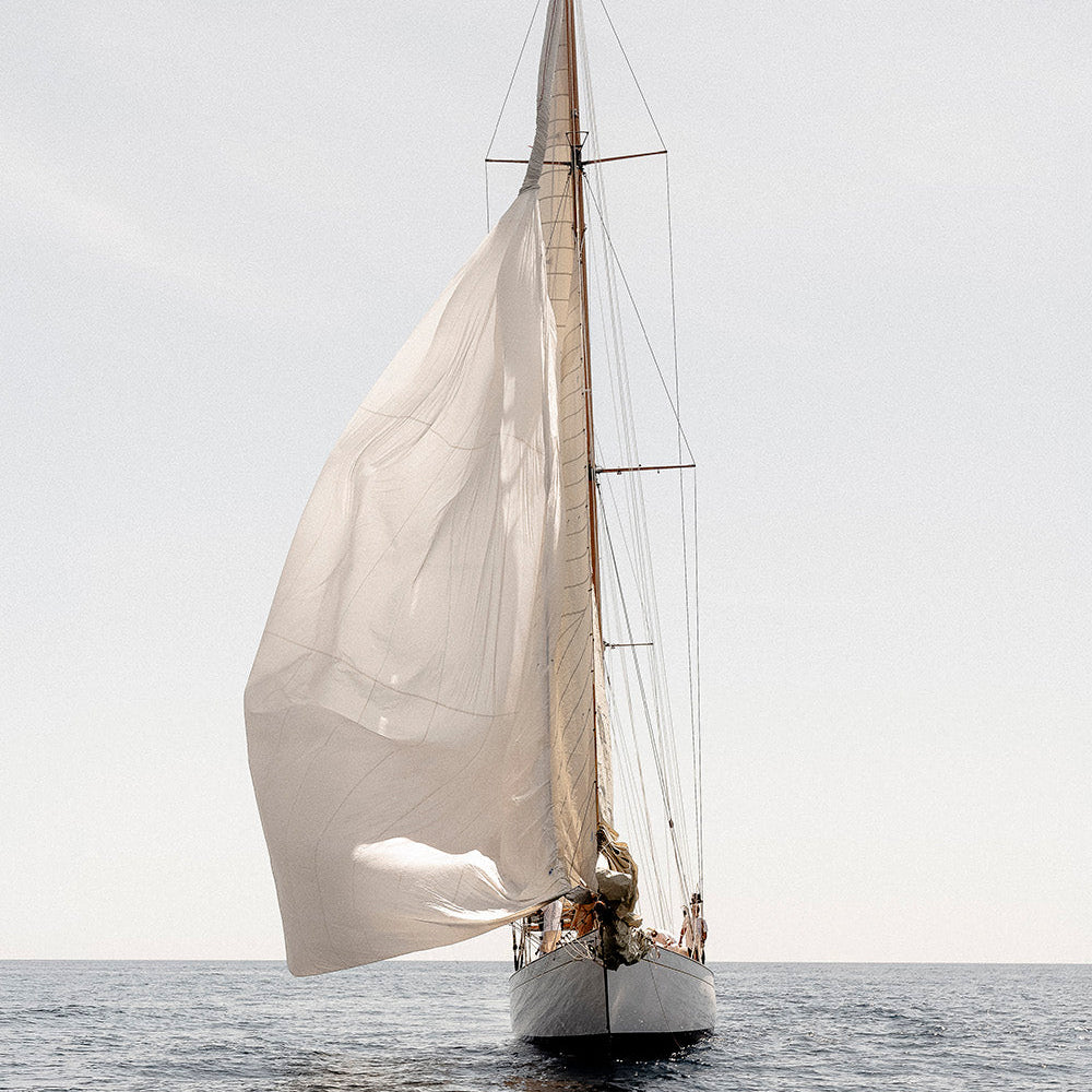 A white sailboat with large white sails sailing on calm blue-gray water under a pale gray sky.
