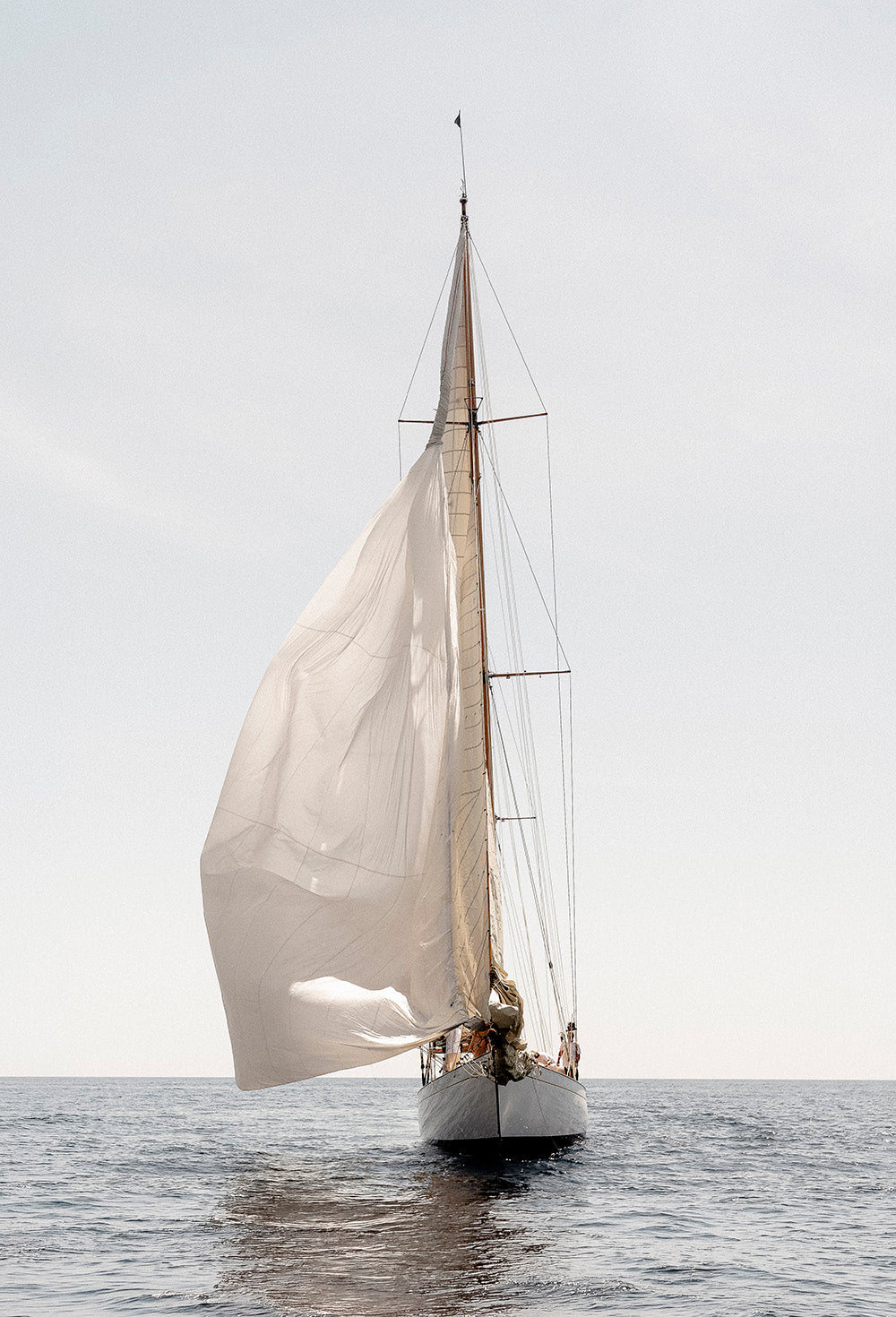 A white sailboat with large white sails sailing on calm blue-gray water under a pale gray sky.