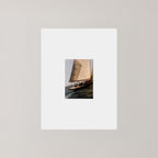 A vertical art print featuring a photograph of a sailboat on dark water, surrounded by a wide white border and set against a neutral light gray background.
