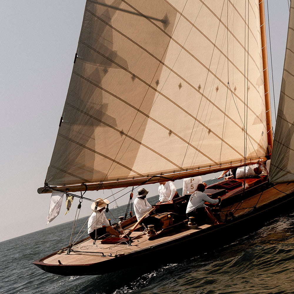 A traditional wooden sailing yacht with large beige sails cuts through the ocean waves under a clear sky, with crew members visible on board.
