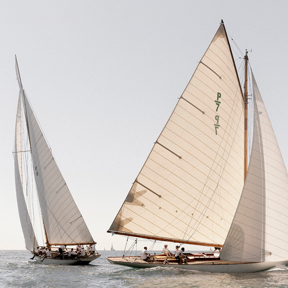 Three classic white-sailed yachts race across calm blue-gray water under a pale overcast sky. The largest yacht in the foreground has its mainsail fully unfurled.