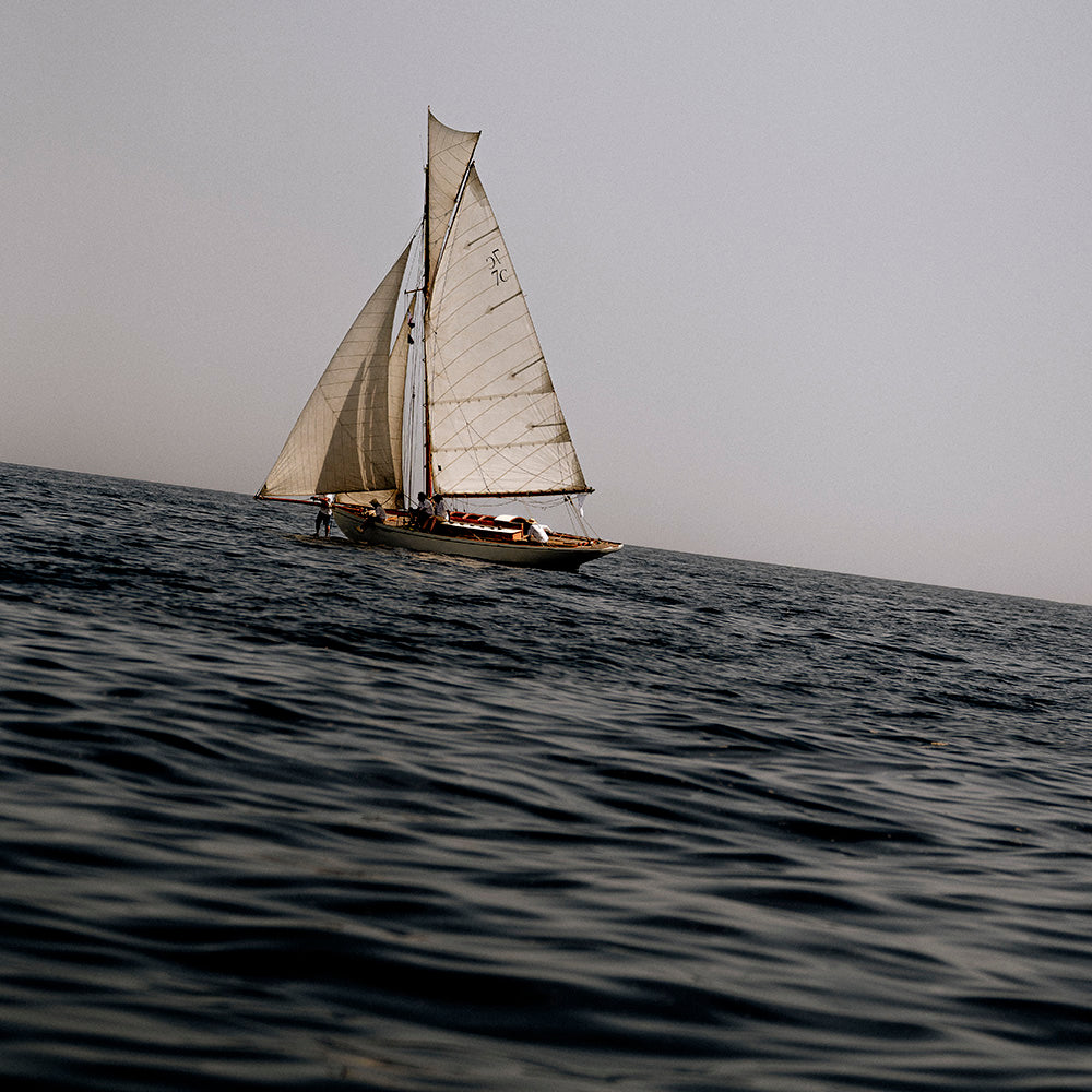 A small sailboat with white sails traveling across dark blue ocean waters under a flat, overcast gray sky.