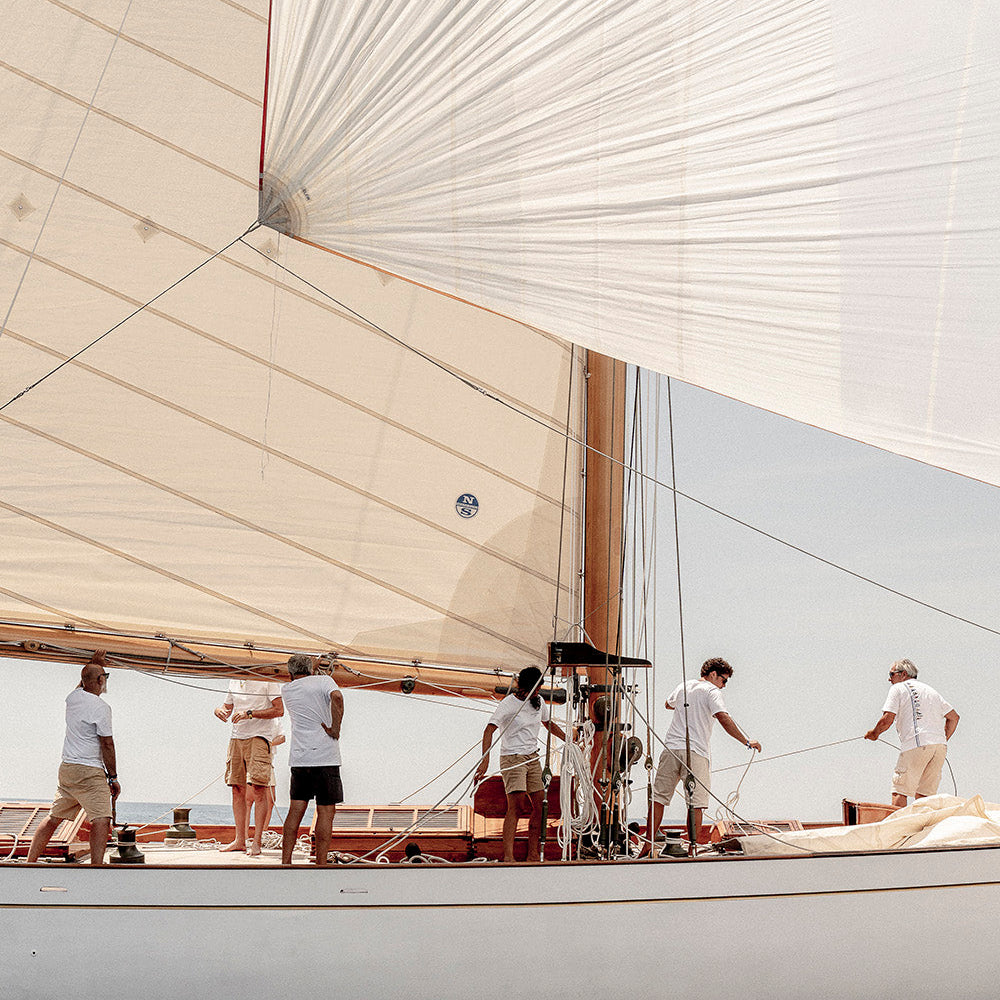 A large white sailboat on the water with several people standing on deck under a bright sky, with the sail filling much of the frame.