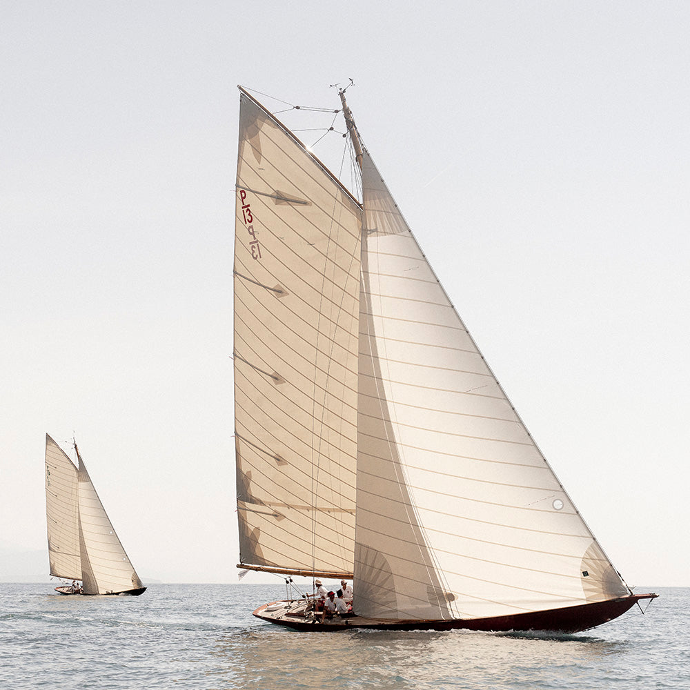 A large traditional sailboat with tall cream-colored sails sailing on calm water, with a smaller similar sailboat visible in the background under a pale sky.