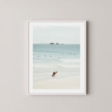 Wategos Calm art print of a surfer walking with a surfboard on the beach in Byron Bay, Australia, with the ocean and other surfers in the background, displayed on a wall in a white frame.