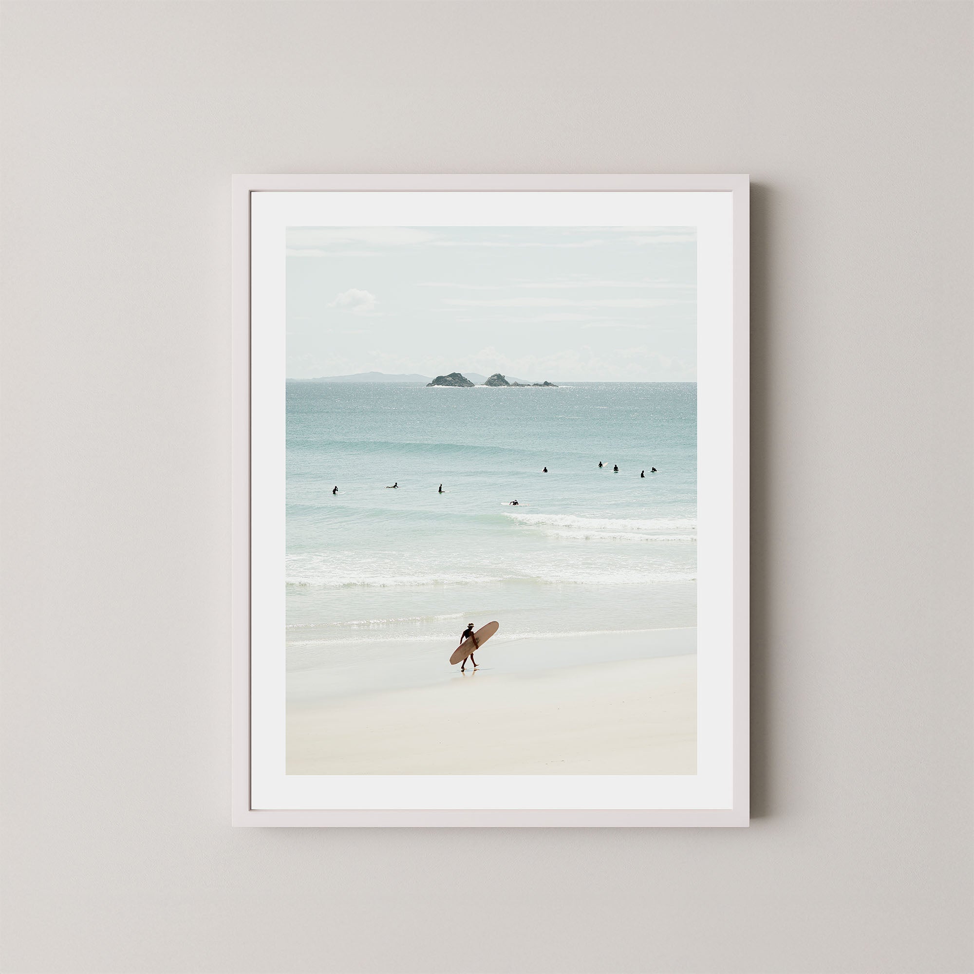 Wategos Calm art print of a surfer walking with a surfboard on the beach in Byron Bay, Australia, with the ocean and other surfers in the background, displayed on a wall in a white frame.