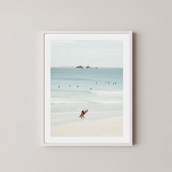 Wategos Calm art print of a surfer walking with a surfboard on the beach in Byron Bay, Australia, with the ocean and other surfers in the background, displayed on a wall in a white frame.