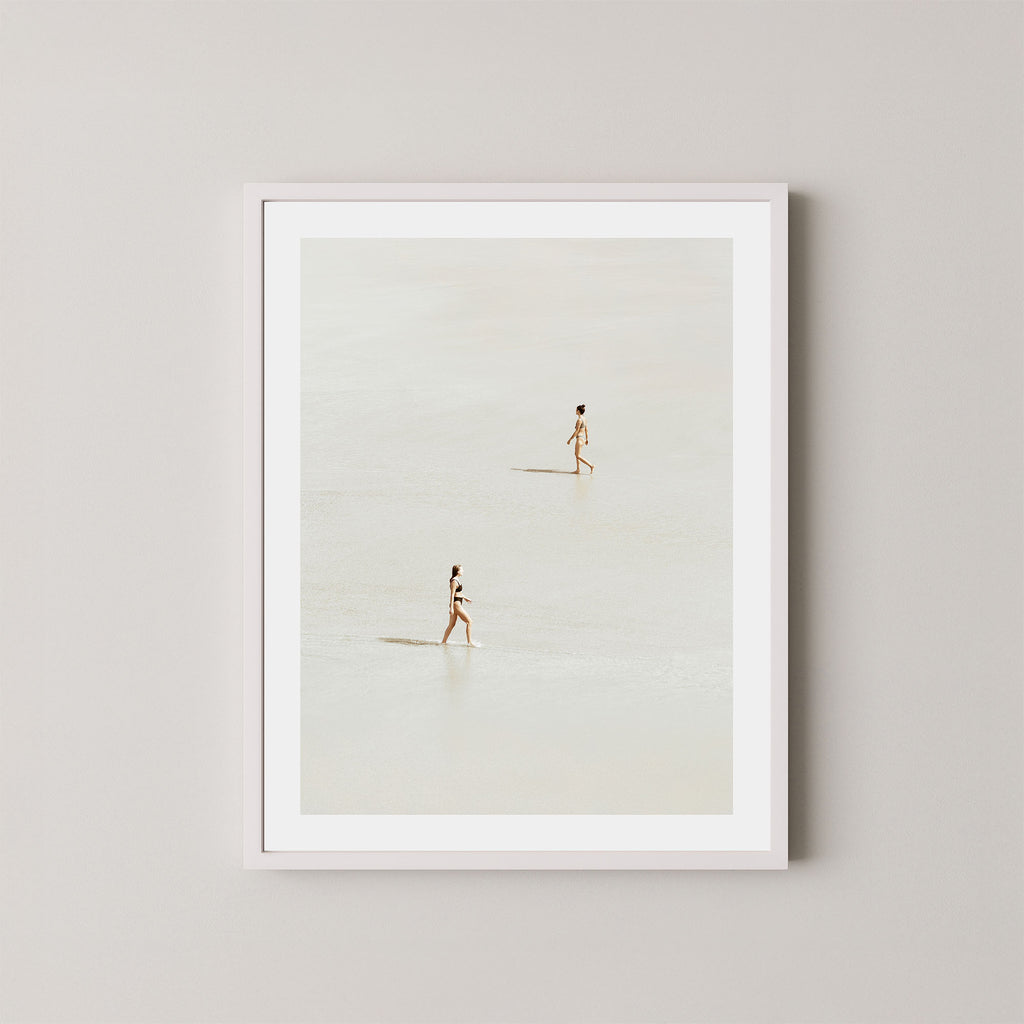 Beach Crossing art print featuring two women walking in opposite directions on a light, sandy beach at low tide in Byron Bay, Australia, displayed on a wall in a white frame.
