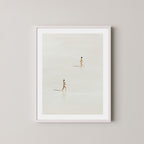 Beach Crossing art print featuring two women walking in opposite directions on a light, sandy beach at low tide in Byron Bay, Australia, displayed on a wall in a white frame.